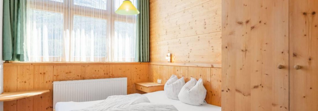 Ein gemütliches Schlafzimmer aus Holz mit einem großen Fenster und viel Tageslicht. Das Bett ist mit einer weißen Decke und Kissen ausgestattet. - Garni Nadine - Sölden
