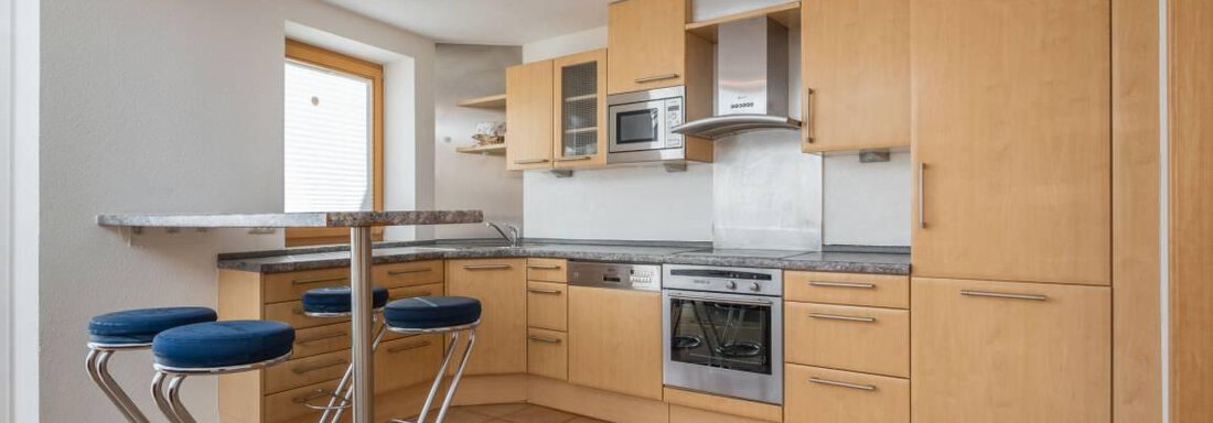 Eine moderne Küche mit Holzschränken und einer eleganten Bar mit blauen Hockern. Die schlichte Ausstattung sorgt für ein offenes und freundliches Ambiente. - Garni Nadine - Sölden