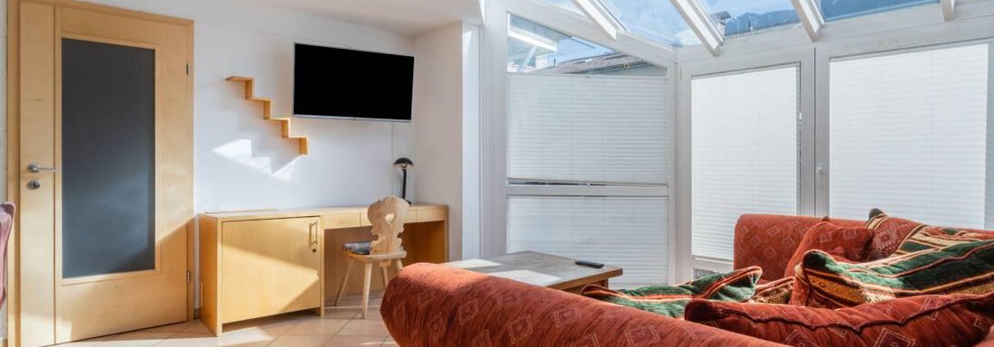 Ein heller Wohnraum mit einem großen Fenster und einem roten Sofa. Zu sehen sind auch ein Holzregal und ein Fernseher an der Wand. - Garni Nadine - Sölden