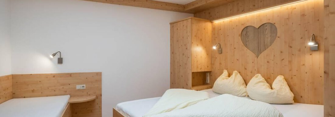 Ein gemütliches Schlafzimmer mit Holzmöbeln und einem großen Doppelbett. An der Wand ist ein herzförmiges Fenster, das für eine charmante Atmosphäre sorgt. - Garni Nadine - Sölden