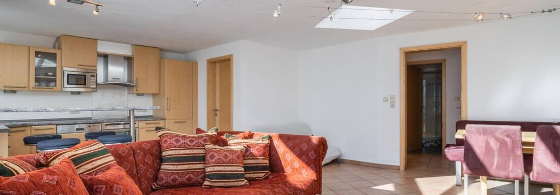 Ein helles Wohnzimmer mit einem roten Sofa und einem Holztisch. Die offene Küche ist gut ausgestattet und es gibt große Fenster, die viel Licht hereinscheinen lassen. - Garni Nadine - Sölden