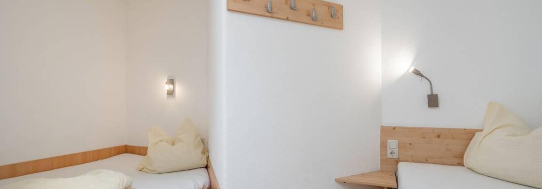 Ein einfaches Schlafzimmer mit zwei Einzelbetten. Die Wände sind hell und es gibt eine Holzverkleidung sowie einige Wandhaken. - Garni Nadine - Sölden