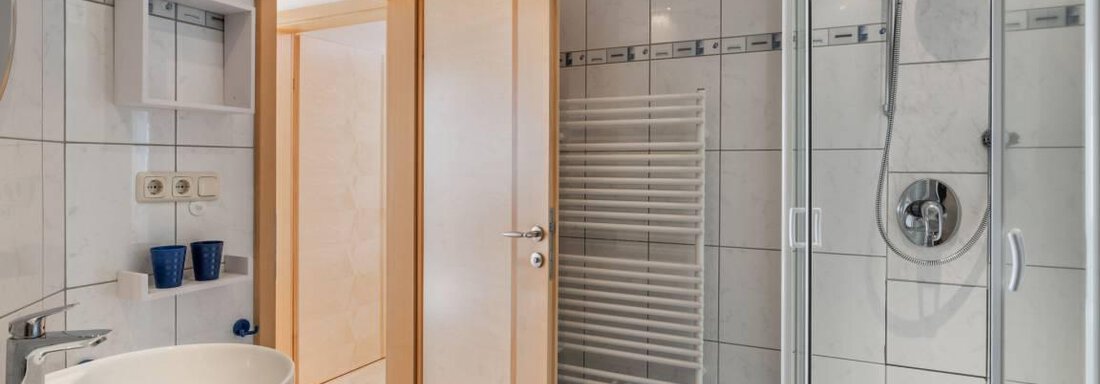 Ein modernes Badezimmer mit einer Dusche, einem Waschbecken und einem Handtuchwärmer. Die Wände sind hell gefliest und es gibt eine offene Tür zu einem weiteren Raum. - Garni Nadine - Sölden