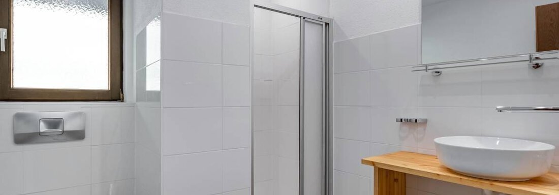 Ein modernes Badezimmer mit weißen Fliesen und einer Dusche. Ein Waschbecken auf einem Holzregal und ein Fenster sorgen für helle Lichtverhältnisse. - Garni Nadine - Sölden