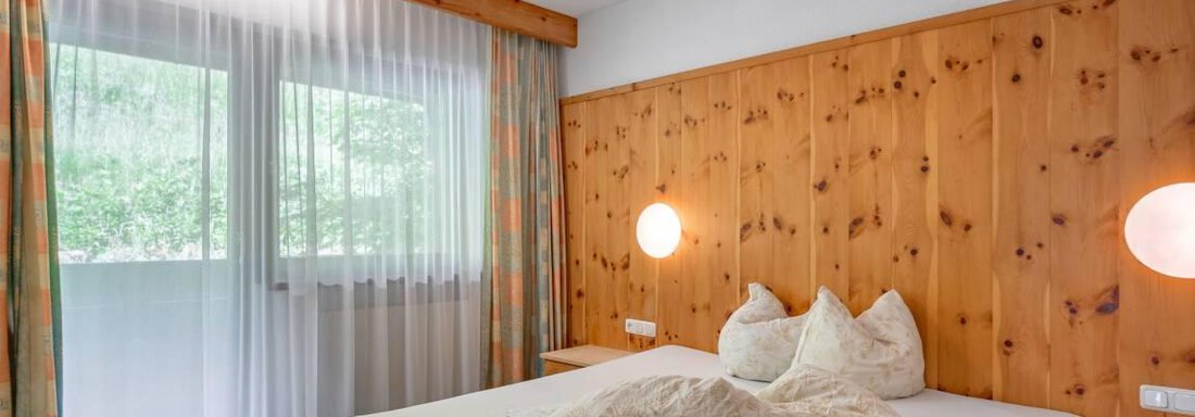 Ein gemütliches Schlafzimmer mit einem großen Bett und warmen Holzwänden. Große Fenster bieten viel Tageslicht und Blick nach draußen. - Garni Nadine - Sölden