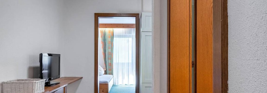 Ein gemütlicher Flur mit Holzdecke und einem Fernseher. Im Hintergrund ist ein heller Raum mit einem Bett und einem Balkon sichtbar. - Garni Nadine - Sölden