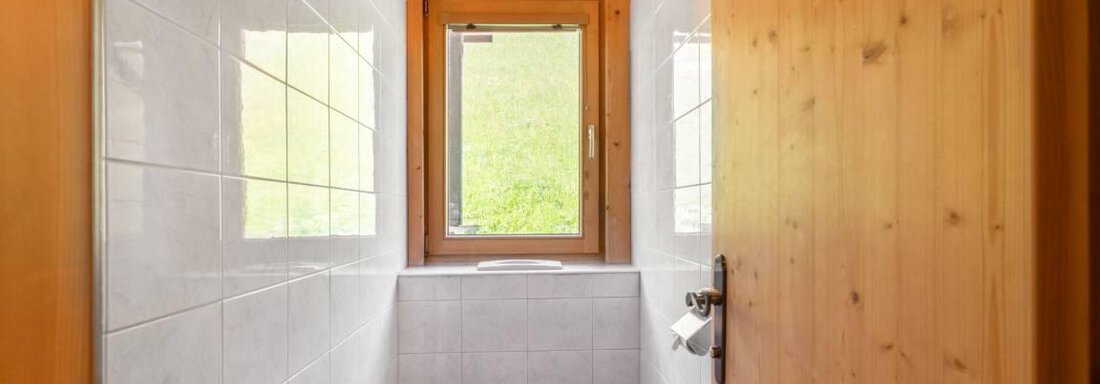 Ein modernes Badezimmer mit weißen Fliesen und einem Fenster. Die Holztür und -rahmen verleihen dem Raum eine warme Atmosphäre. - Garni Nadine - Sölden