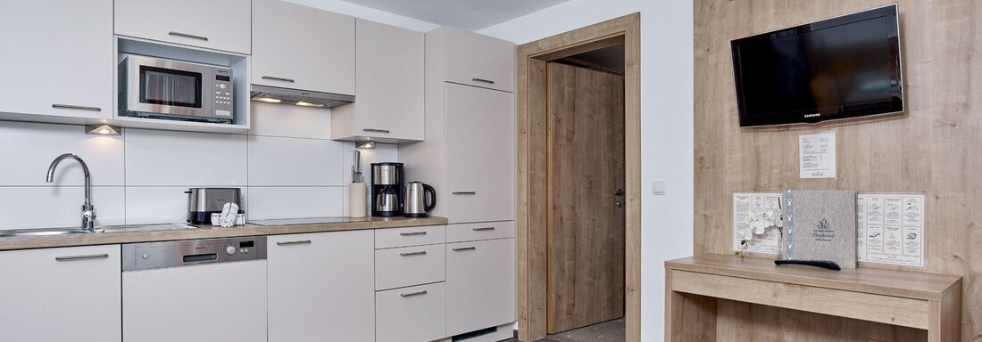 Eine moderne Küche mit hellen Schränken und einer Mikrowelle. An der Wand befindet sich ein Fernseher und ein Tisch aus Holz. - Gästehaus Bergkristall - Sölden