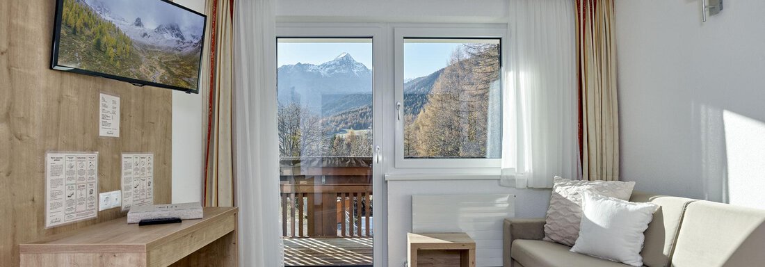 Ein helles Wohnzimmer mit einem Sofa und einem Fernseher. Die Fenster bieten einen Blick auf die Berge. - Gästehaus Bergkristall - Sölden