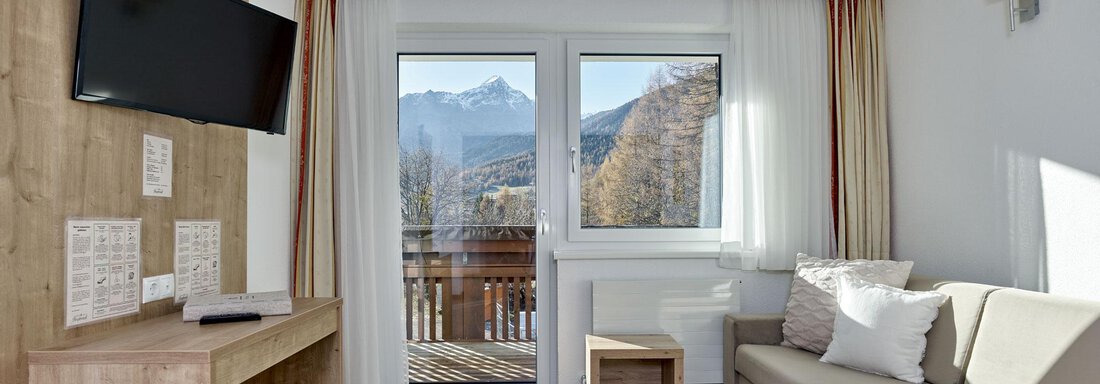 Ein gemütliches Wohnzimmer mit einem modernen Sofa und einem Fernseher. Große Fenster bieten einen Blick auf die Berge und viel Tageslicht. - Gästehaus Bergkristall - Sölden