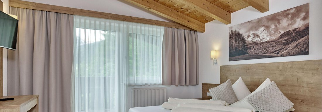 A modern hotel room with wooden ceilings and large windows. There is a double bed with pillows and a small seating area. - Gästehaus Bergkristall - Sölden