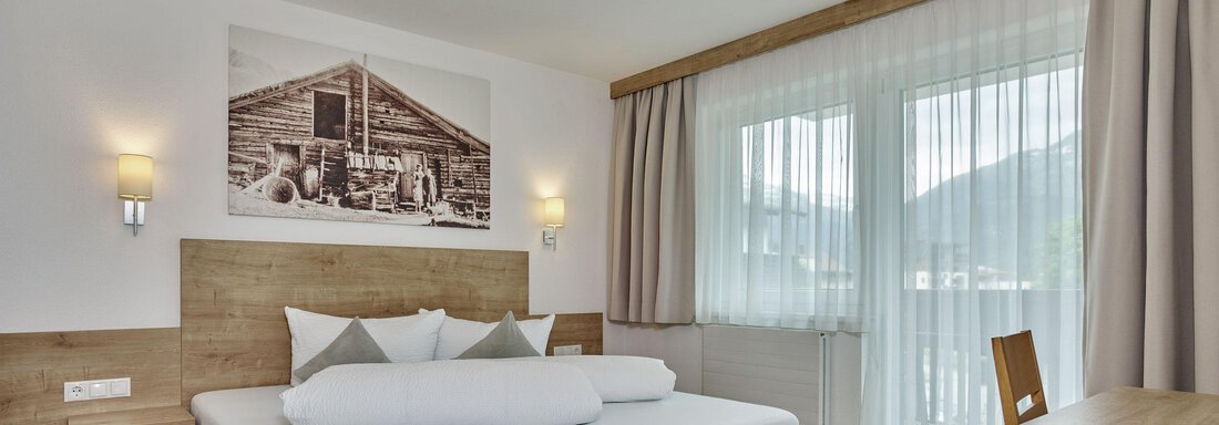 Ein modernes Schlafzimmer mit einem Holzrahmenbett und sanften Farben. Große Fenster lassen viel Licht herein und bieten einen Blick nach draußen. - Gästehaus Bergkristall - Sölden