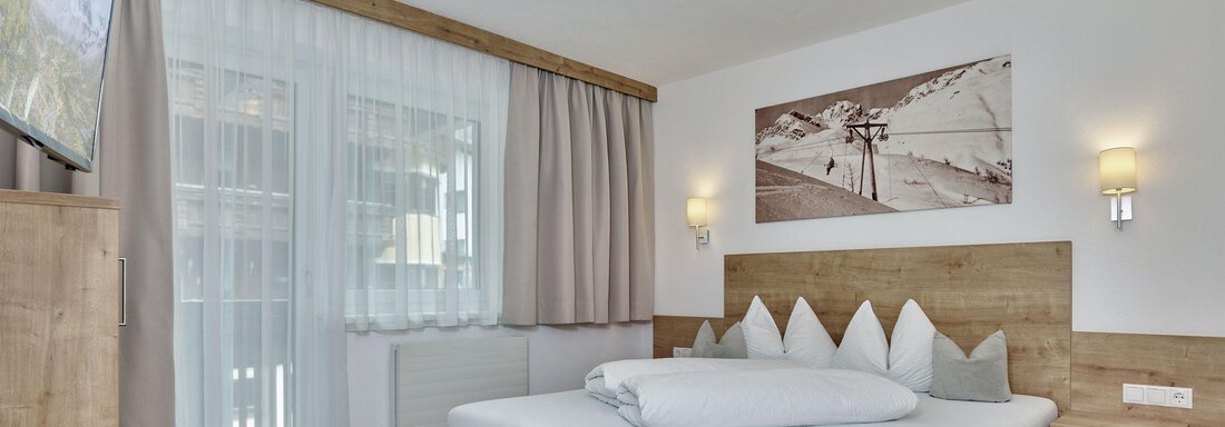 Ein modernes Schlafzimmer mit einem großen Bett und Holzmöbeln. Der Raum hat eine helle Atmosphäre und einen Blick auf die Natur. - Gästehaus Bergkristall - Sölden