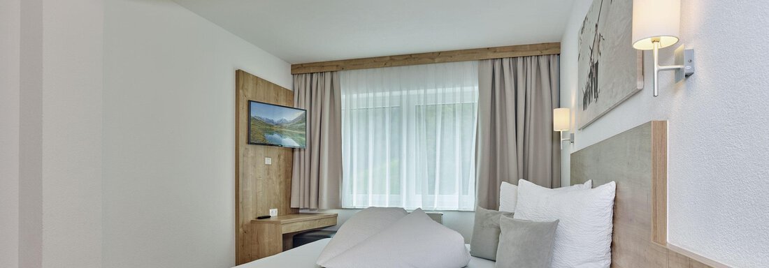 Ein modernes Schlafzimmer mit einem großen Bett und hellen, freundlichen Farben. Es gibt Fenster mit Vorhängen und eine kleine Fernsehecke. - Gästehaus Bergkristall - Sölden