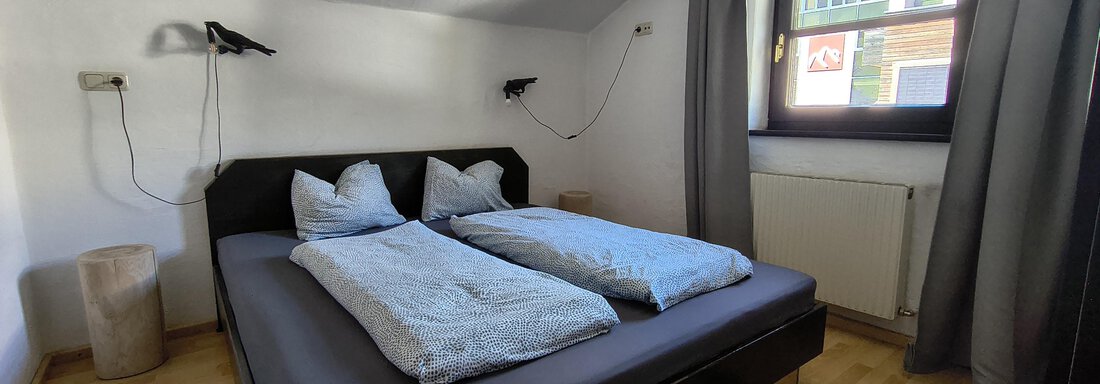 A cozy bedroom with a double bed and two pillows. The room has wooden floors and a window with curtains. - Gästehaus Hermann Arnold - Sölden