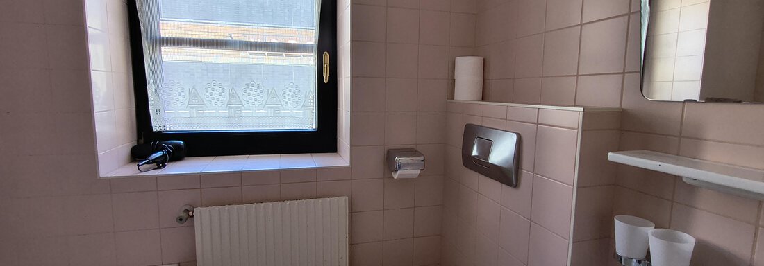 A bright bathroom with pink tiles. There is a bathtub, a toilet, and a sink under a large window. - Gästehaus Hermann Arnold - Sölden