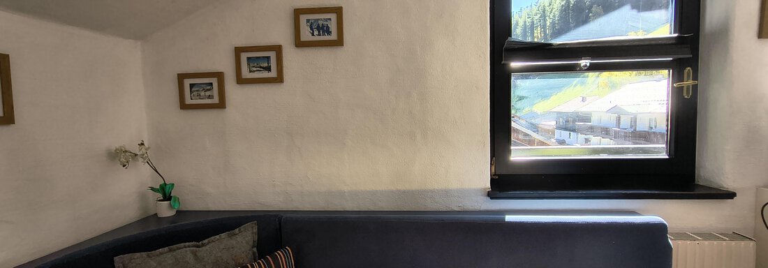 A cozy room with a blue seating area and a window that offers a view of the mountains. Some picture frames hang on the wall. - Gästehaus Hermann Arnold - Sölden