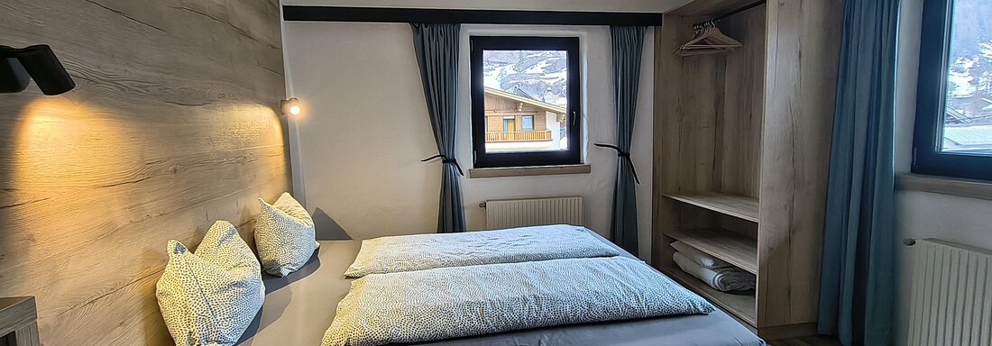 The image shows a modern bedroom with a large bed and bright wooden walls. Through the window, there is a view of the surrounding landscape. - Gästehaus Hermann Arnold - Sölden