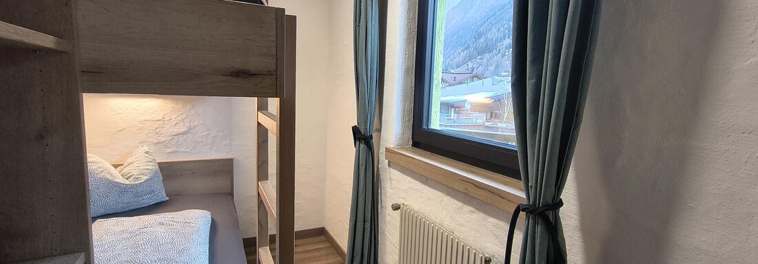 A simple room with a bunk bed and a large window. The walls are bright and the curtains are blue. - Gästehaus Hermann Arnold - Sölden