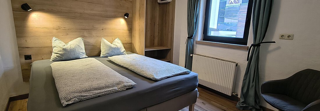 A modern bedroom with a double bed and wood decor. There is an armchair and a window that lets in daylight. - Gästehaus Hermann Arnold - Sölden