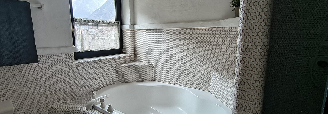 A modern bathroom with an oval bathtub. The window lets in daylight and the walls are painted in light colors. - Gästehaus Hermann Arnold - Sölden
