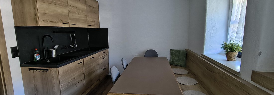 A modern kitchen with wooden cabinets and a black countertop. A large dining table with chairs and a cozy bench can also be seen. - Gästehaus Hermann Arnold - Sölden