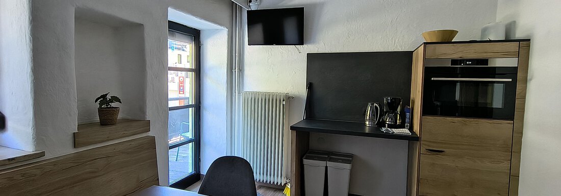 A modern kitchen with a wooden table and a chair. In the background, there is a stove, a microwave, and a window with a view outside. - Gästehaus Hermann Arnold - Sölden