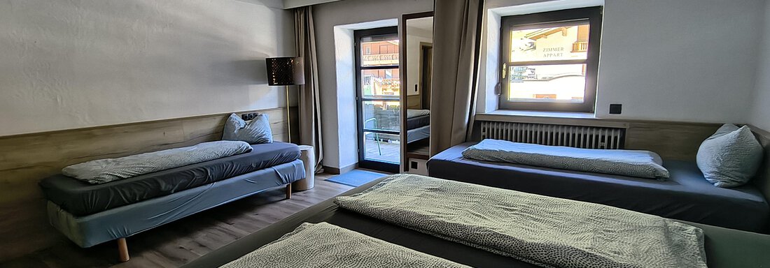 A modern room with three beds and a window that leads to a balcony. The decor is simply and cozily designed. - Gästehaus Hermann Arnold - Sölden
