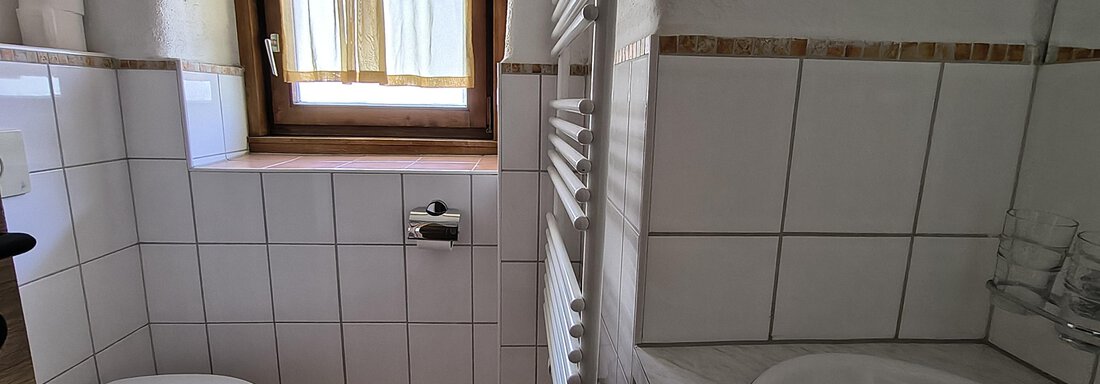 A small bathroom with a toilet, sink, and a bathtub. There is a window with curtains and bright tiles. - Gästehaus Hermann Arnold - Sölden