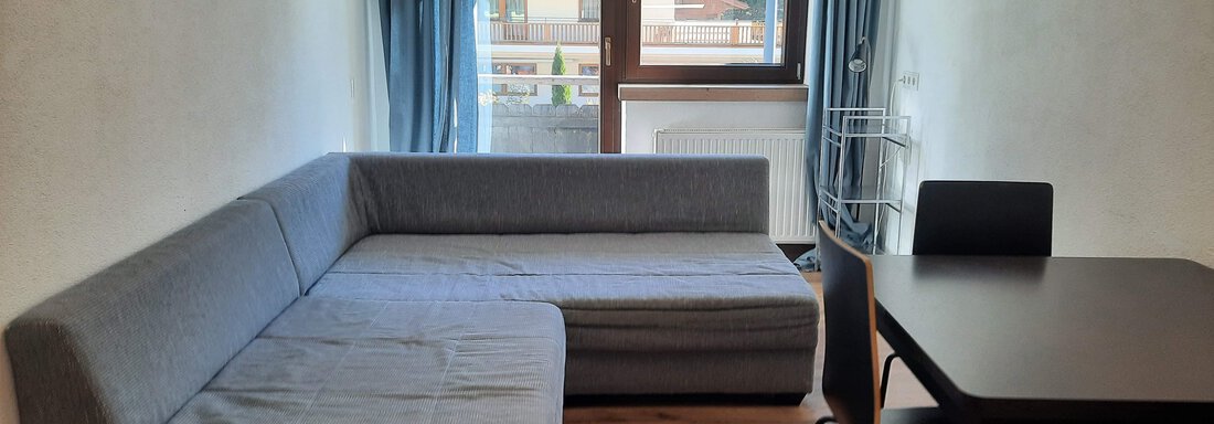 A cozy living space with a grey sofa and a table with black chairs. The large windows let in plenty of daylight and offer a view outside. - Gästehaus Hermann Arnold - Sölden