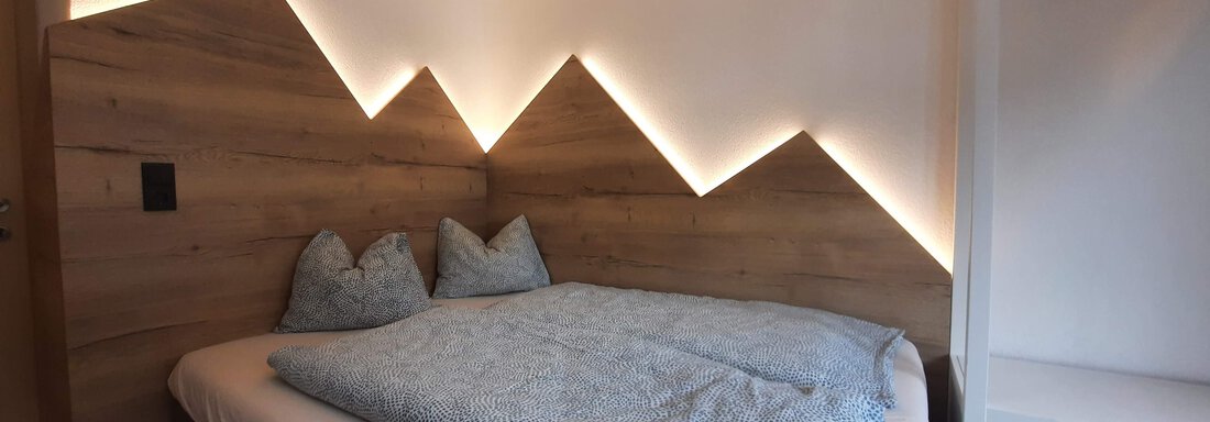 A modern bedroom with a bed and a stylish wooden wall paneling. The soft lighting above the headboard creates a cozy atmosphere. - Gästehaus Hermann Arnold - Sölden