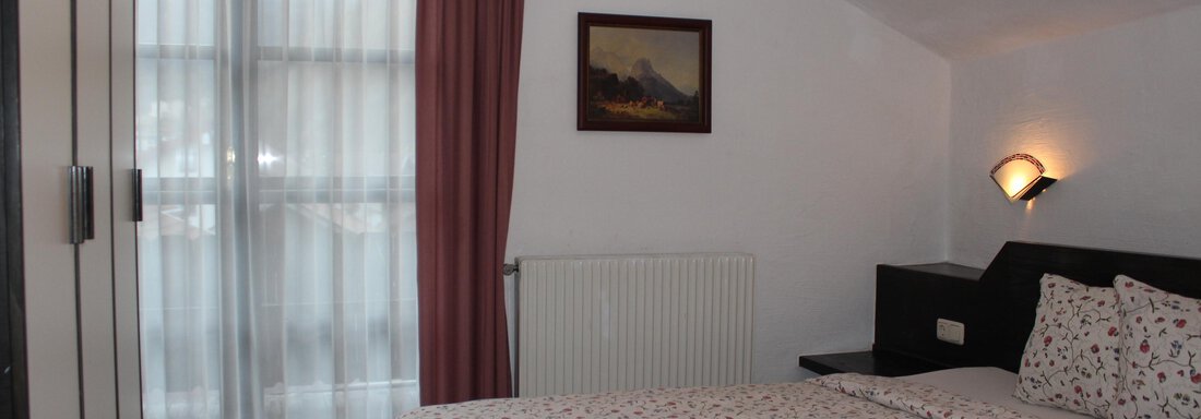 A cozy bedroom with a double bed and a window. The walls are light, and curtains as well as a small painting create a warm atmosphere. - Gästehaus Hermann Arnold - Sölden
