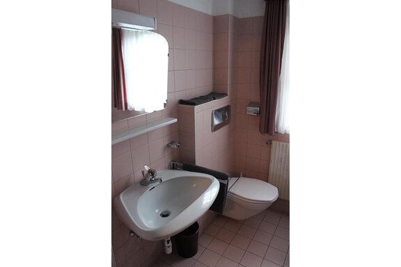 A simple bathroom with pink tiles. It has a sink, a toilet, and a window with curtains. - Gästehaus Hermann Arnold - Sölden