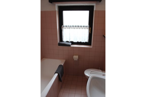A bathroom with a bathtub, a toilet, and a small window. The walls are tiled in gentle shades of pink. - Gästehaus Hermann Arnold - Sölden