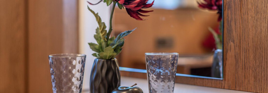 Ein dekoratives Tablett mit einem Blumenarrangement und zwei Gläsern. Im Hintergrund spiegelt sich ein Spiegel. - Gästehaus Maier - Sölden