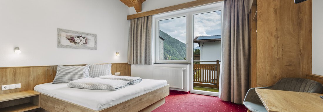 Ein helles Zimmer mit Holzdecke und rotem Teppich. Es enthält ein Bett, einen Tisch und einen Balkon mit Blick nach draußen. - Gasthof Granstein - Sölden