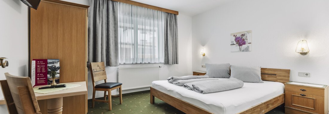 Ein einfaches und gemütliches Zimmer mit einem Doppelbett und einem Fernseher. Es gibt einen Schreibtisch und Stühle sowie eine helle Fensterfront. - Gasthof Granstein - Sölden