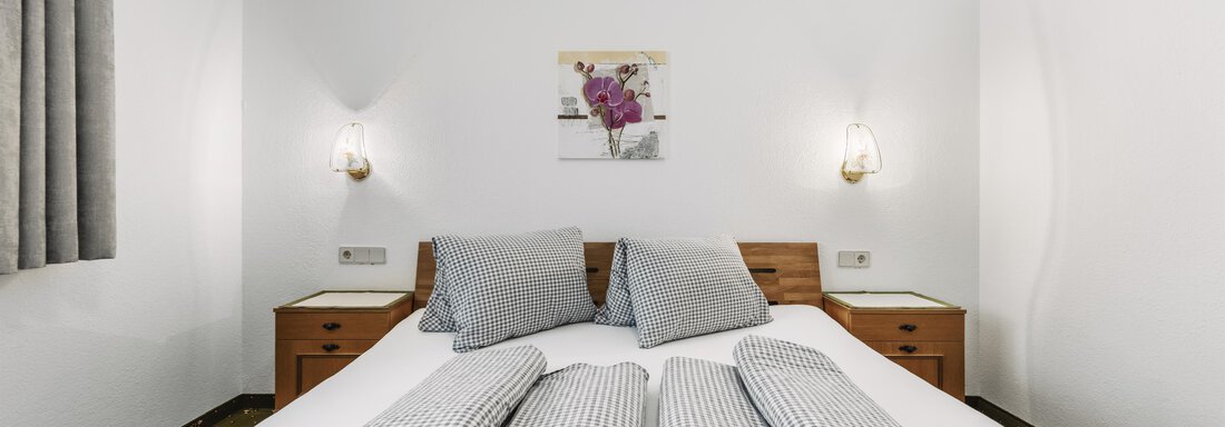 Ein gemütliches Schlafzimmer mit einem großen Bett und gestreiften Kissen. An den Wänden hängen Leuchten und ein kleines Kunstwerk. - Gasthof Granstein - Sölden