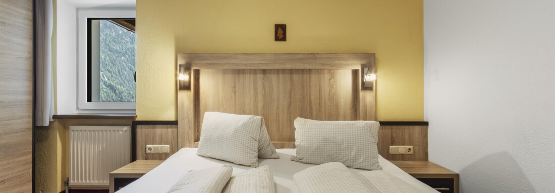 Ein gemütliches Schlafzimmer mit einem Doppelbett und hellem Holzinterieur. Die Wände sind in warmem Gelb gestrichen und ein Fenster lässt viel Tageslicht herein. - Gasthof Granstein - Sölden