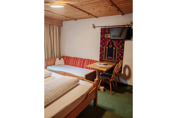 Ein gemütliches Zimmer mit zwei Betten und einer Sitzecke. An der Wand hängt ein dekorativer Teppich, und ein Fernseher ist ebenfalls vorhanden. - Haus der Bergfreunde - Sölden