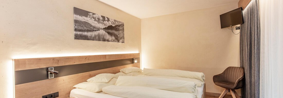 Zimmer Top 1 Ein modernes Schlafzimmer mit einem Doppelbett und hellem Holzdesign. An der Wand hängt ein Bild einer Landschaft, und es gibt einen bequemen Stuhl und ein Fenster mit Gardinen. - Haus Manfred - Sölden