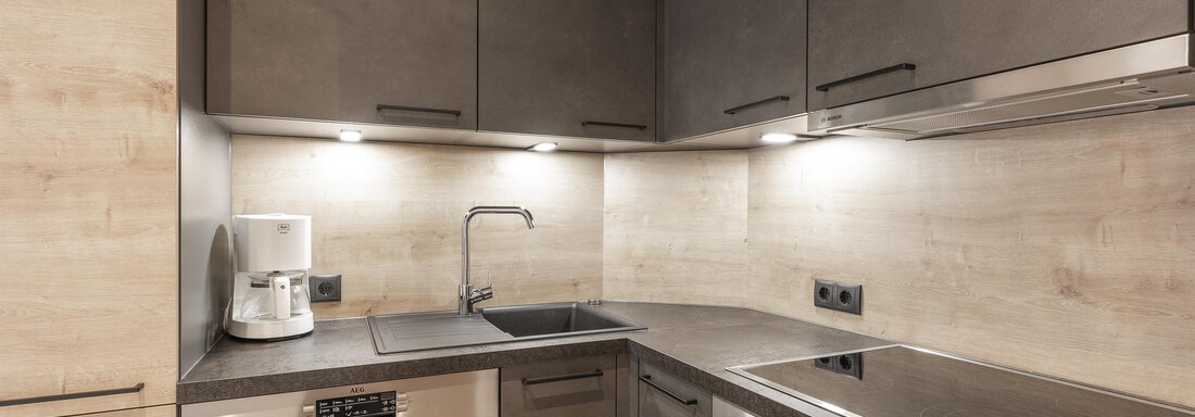 Eine moderne Küche mit dunklen Schränken und einer hellen Arbeitsplatte. Die Ausstattung umfasst eine Spüle, einen Geschirrspüler und ein Kochfeld. - Haus Manfred - Sölden