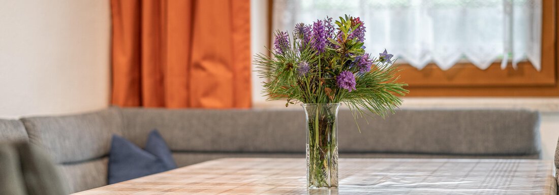 A beautifully set table with a vase full of colorful flowers. In the background, you can see a cozy seating area with orange curtains. - Haus Nicola - Sölden