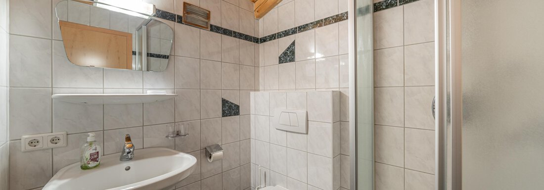 A modern bathroom with a shower and sink. The walls are tiled, and there is a wooden beam ceiling. - Haus Nicola - Sölden