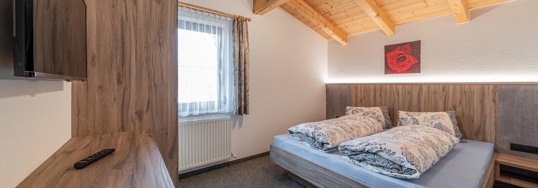 A modern bedroom with wooden ceilings and a large window. The bed is equipped with two pillows and a blanket, and a television is mounted on the wall. - Haus Nicola - Sölden
