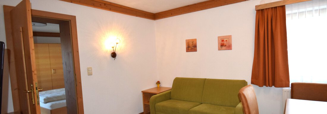 Ein gemütliches Wohnzimmer mit einem grünen Sofa und einem Tisch. Die Wände sind hell, und es gibt Holzverzierungen sowie Bilder. - Haus Romana - Sölden
