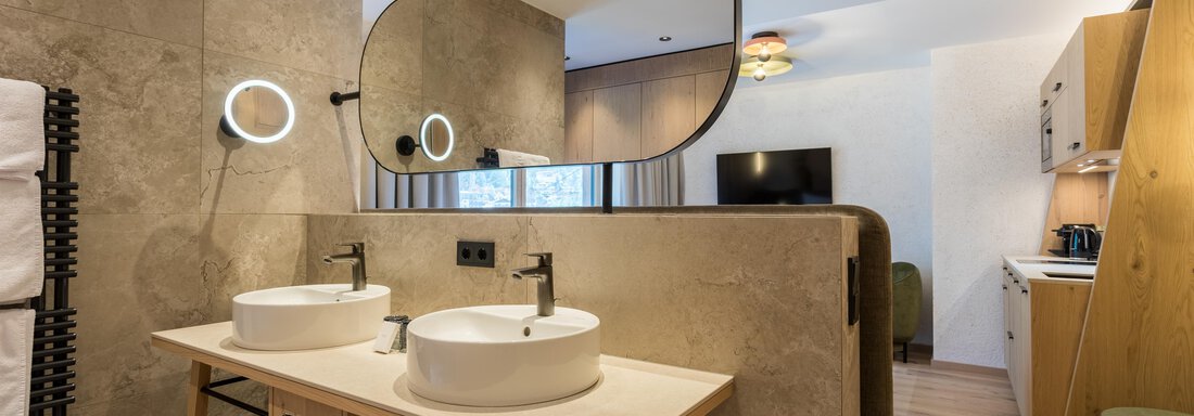 Ein modernes Badezimmer mit zwei Waschbecken und einem großen Spiegel. Die Wände sind in neutralen Farbtönen gehalten und es gibt Holz- und Metallelemente im Design. - Hotel Alpina - Sölden