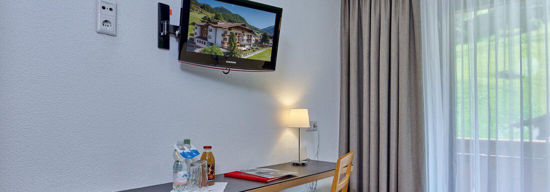 Ein gemütlicher Hotelzimmerbereich mit einem Schreibtisch, einem Fernseher und einem Stuhl. Ein Fenster mit Vorhängen lässt natürliches Licht herein. - Hotel Erhart - Sölden