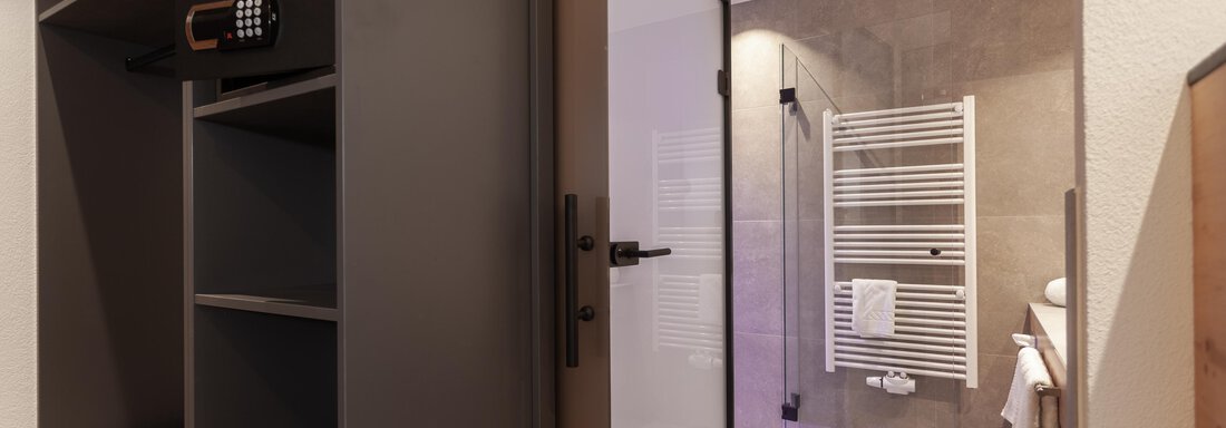 A modern entrance area with an open wardrobe and a bathroom. The shower and the towel warmer are clearly visible. - Hotel Hubertus - Sölden