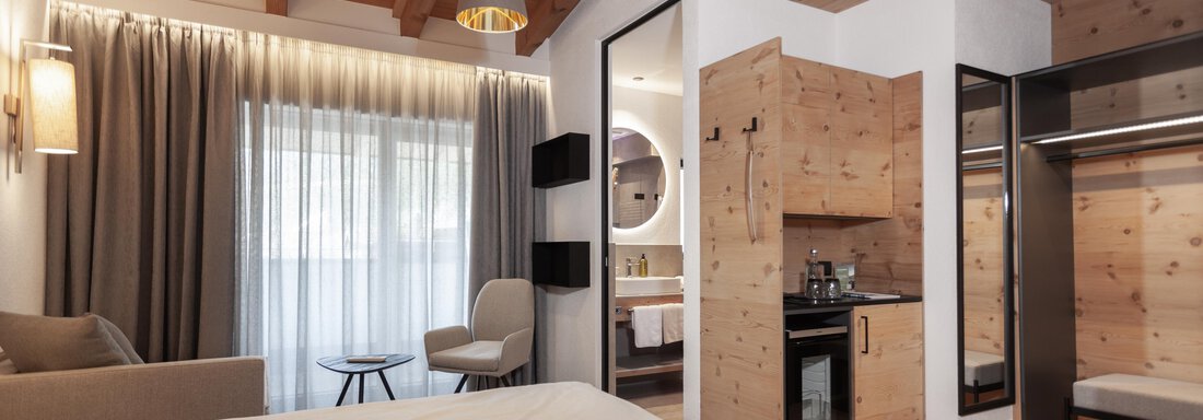 A modern hotel room with two beds, a cozy armchair, and a small kitchenette. Large windows provide plenty of daylight and a pleasant atmosphere. - Hotel Hubertus - Sölden